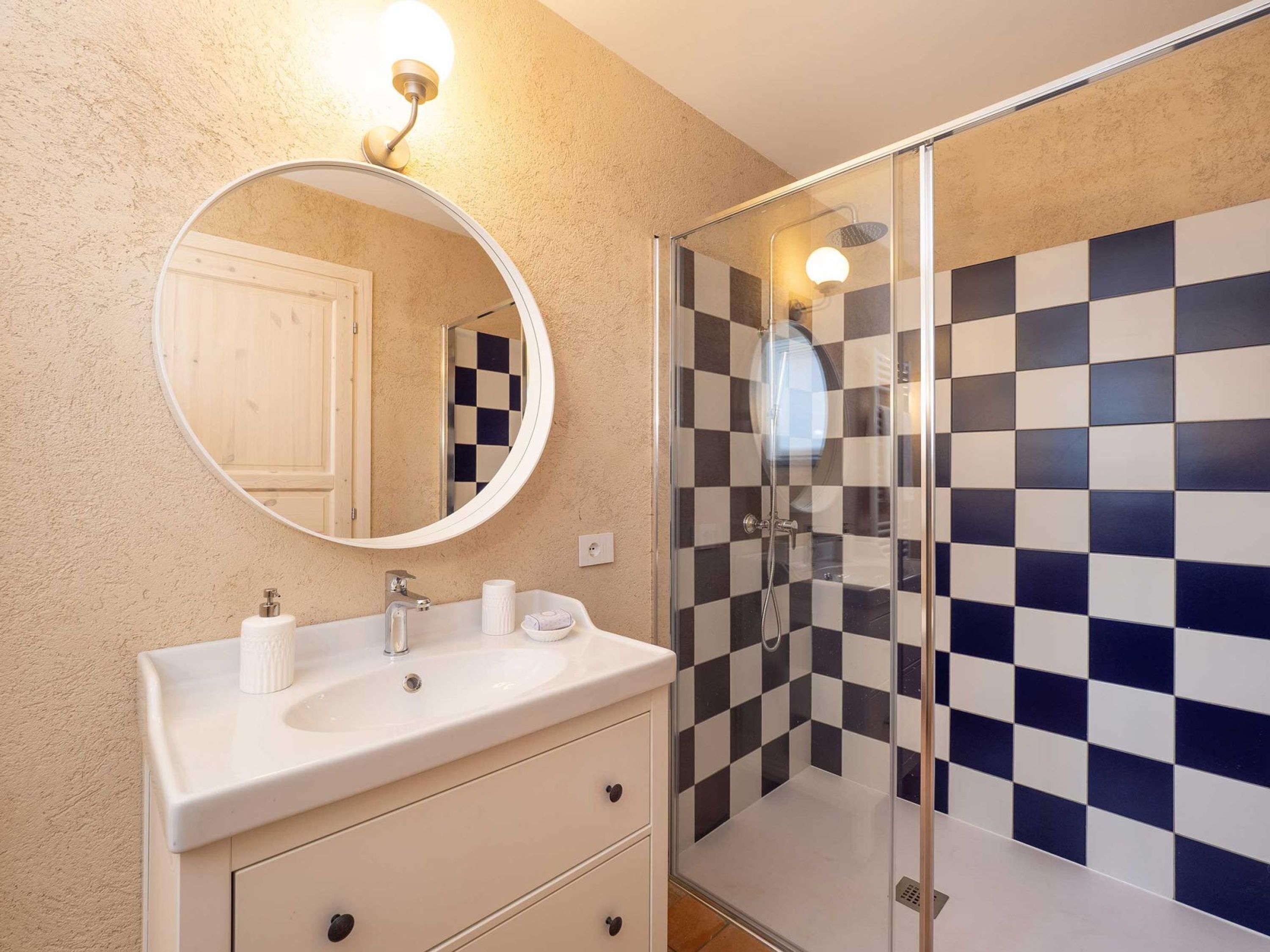 A bathroom with a round mirror, a sink with toiletries, and a shower with checkered tiles.
