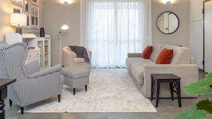 Un salon comprenant un fauteuil à motif pied-de-poule, un canapé blanc avec des coussins orange, une petite table et une grande fenêtre ornée de rideaux légers.