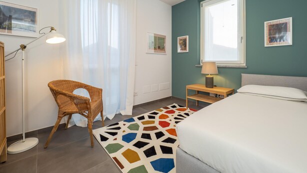 Une chambre à coucher avec un lit, une chaise, une lampe, une fenêtre et des œuvres d’art accrochées aux murs.