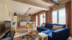 A modern living room with a blue sofa, a small white table, a patterned rug, and a kitchen in the background. - Green Lodge - Apartment 9 - 2 BR 1 Bath (Cressa)