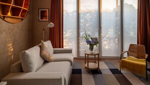 Una sala de estar con un sofá, una mesita de noche con un florero, una silla de mimbre y un ventanal con cortinas.