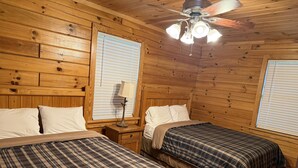 A wooden cabin bedroom with two beds, a ceiling fan, and large windows with blinds.