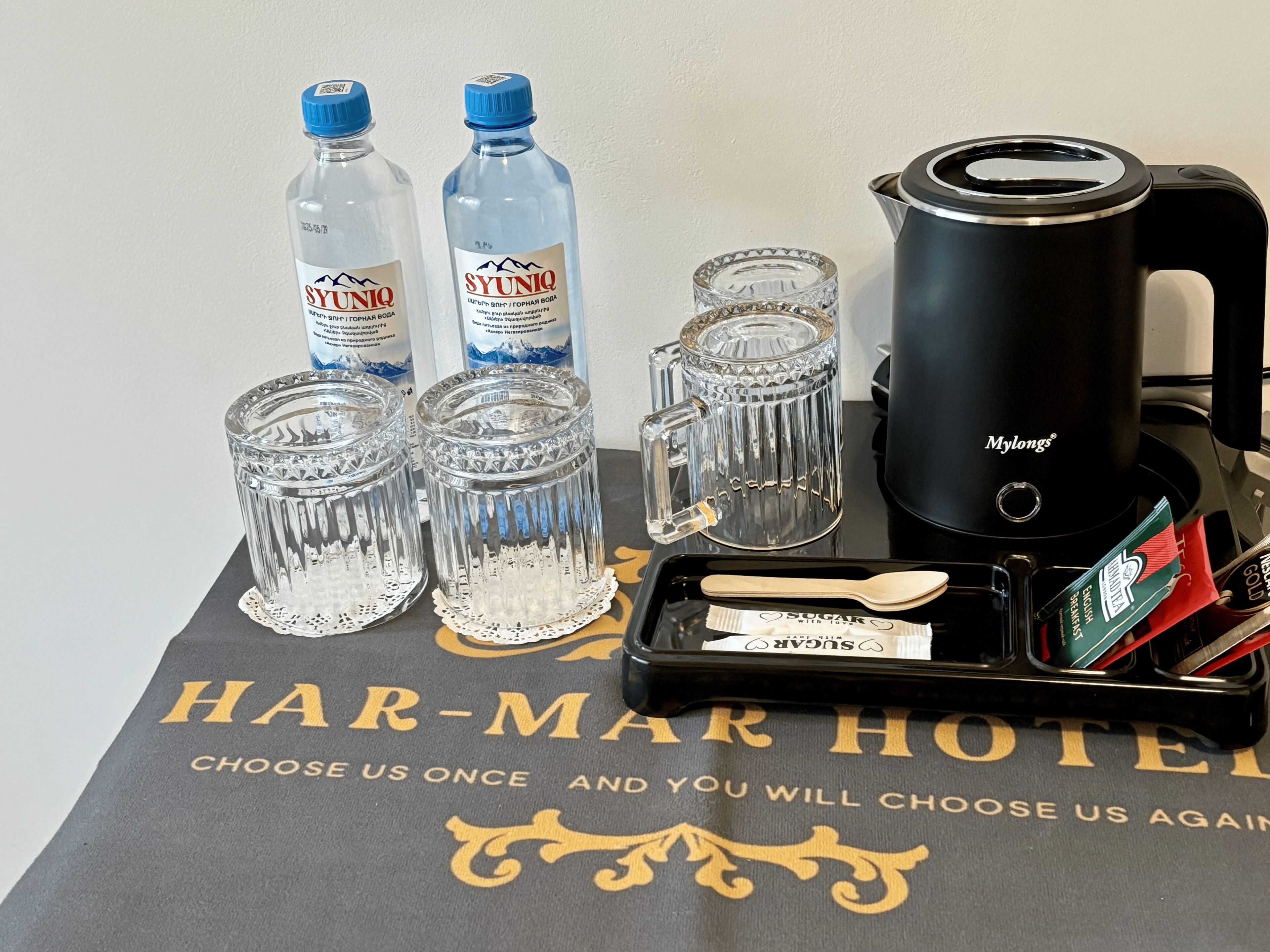 A coffee maker, two water bottles, four glasses, a spoon, and a sugar packet on a table with a hotel towel.