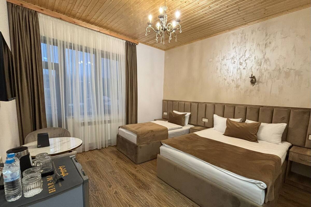 A hotel room with a wooden ceiling, a chandelier, a large bed, a smaller bed, a desk with a chair, and a view through sheer curtains.