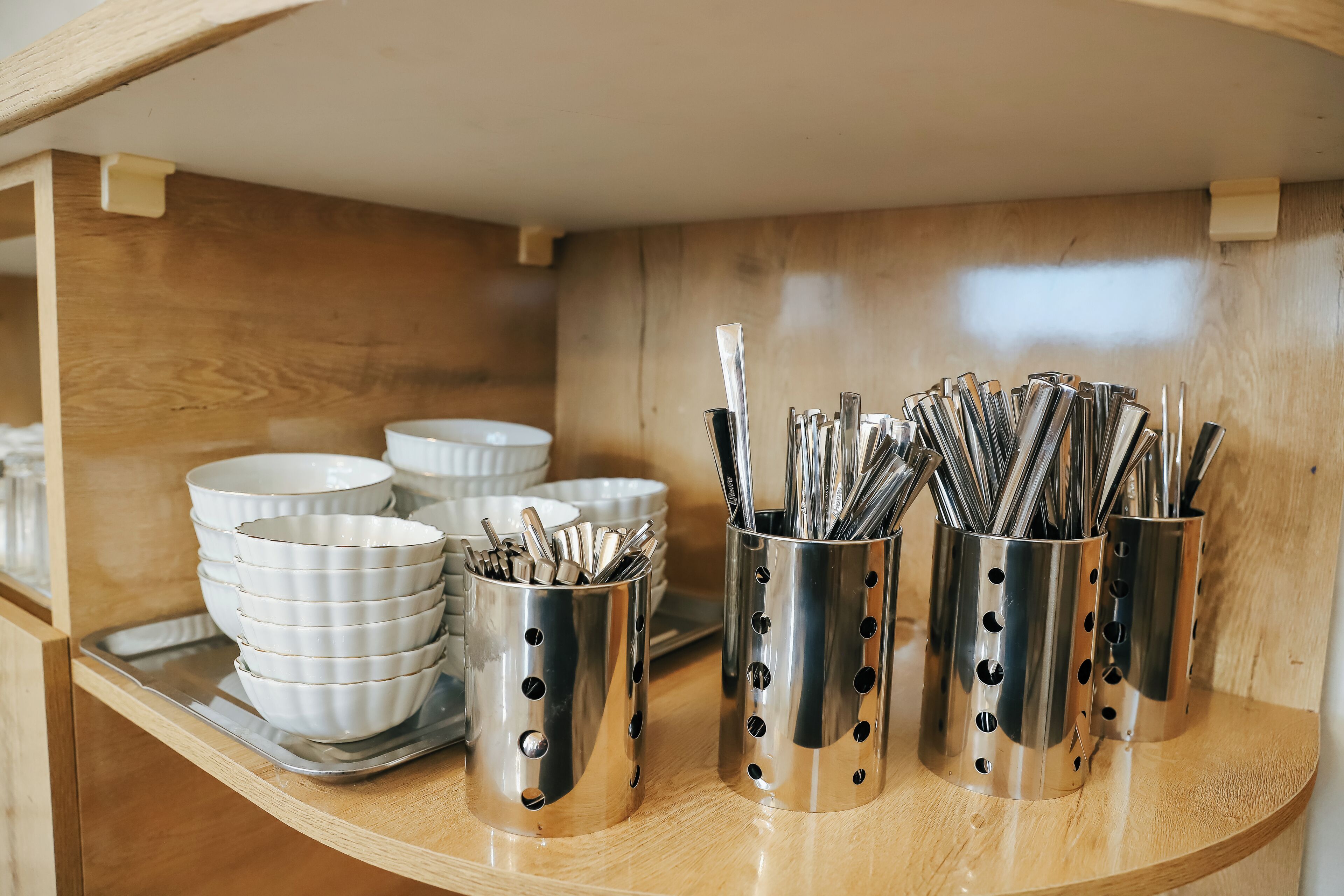Un espacio tipo cocina con utensilios de acero inoxidable y tazones blancos sobre una estantería de madera.
