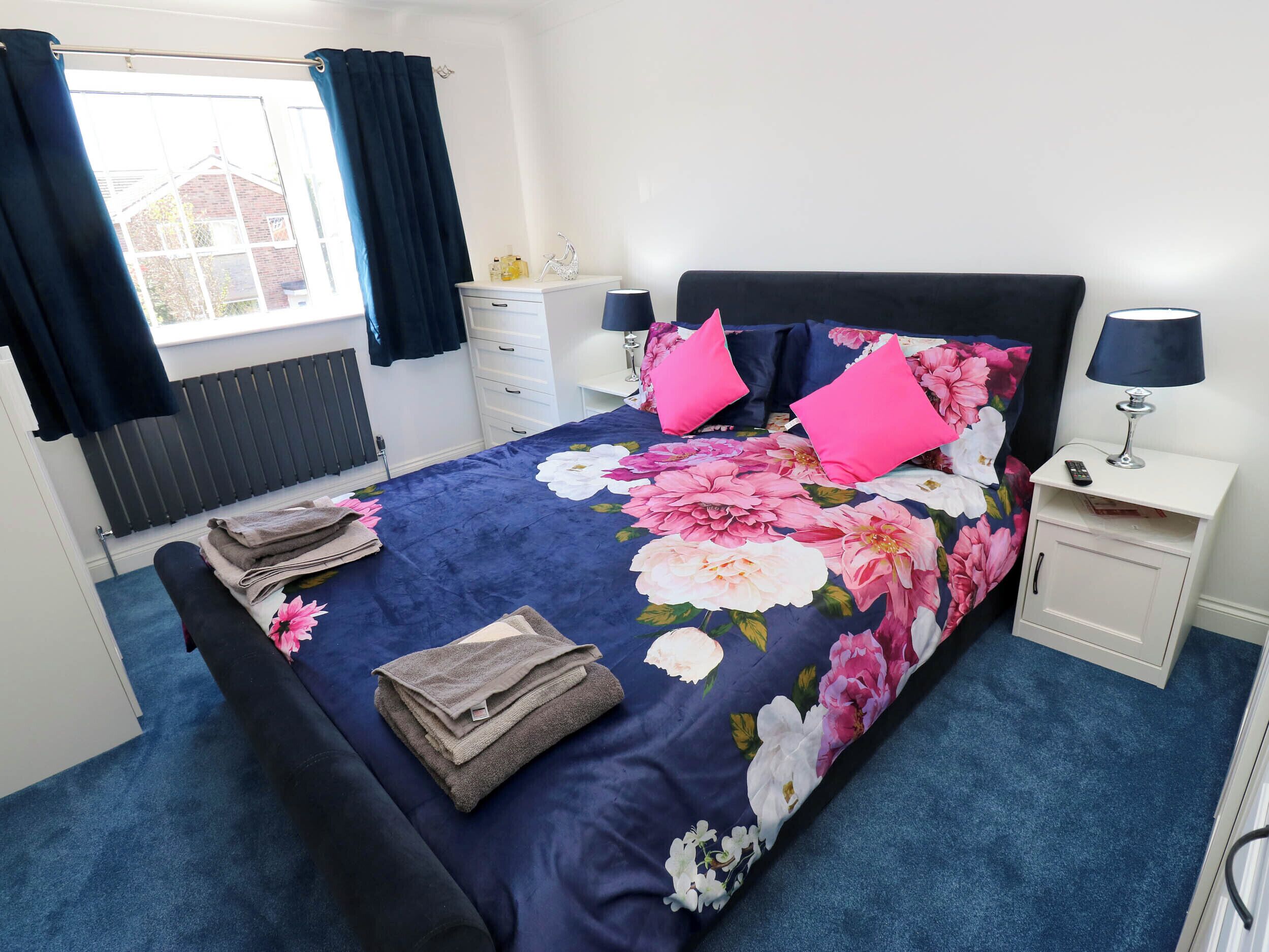 A neatly made bed with a floral duvet, two pillows, and folded towels. There is a bedside table with a lamp and a remote control. A window with curtains is visible, and a radiator is mounted on the wall.