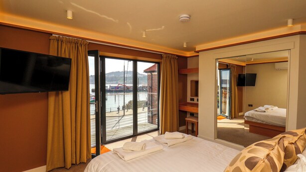 Een hotelkamer met een groot bed, een aan de muur bevestigde televisie en uitzicht op een haven door het raam.