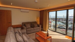 Een woonkamer met een bank, een kleine tafel met een fles en glazen, en uitzicht op een jachthaven door grote ramen.