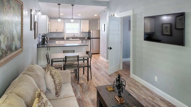 A modern kitchen with white cabinets, a dining area with a wooden table and chairs, and a living room with a beige sofa.
