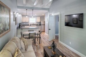 Une cuisine moderne avec des meubles blancs, un coin repas doté d’une table et de chaises en bois, et un salon avec un canapé beige.
