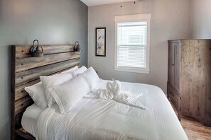 A neatly made bed with white linens and a heart-shaped towel, a rustic wooden headboard, a framed picture on the wall, and a window with blinds. - Historic Bungalow w/ Hot Tub, Walk to St. George (St. Augustine)