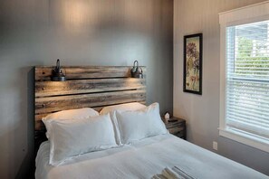 Camera da letto con testiera in legno, biancheria bianca, comodino con lampada e finestra con persiane.