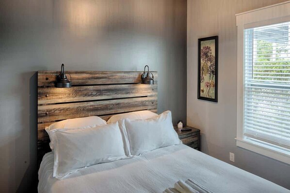 A bedroom with a wooden headboard, white bedding, a bedside table with a lamp, and a window with blinds.