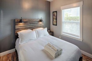 A neatly made bed with white linens and rolled towels, a wooden headboard, and a window with blinds. - Historic St. Augustine Bungalow: Walk to Sites & (St. Augustine)