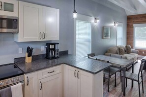 Una cocina moderna con armarios blancos, una estufa integrada y una cafetera. Hay un comedor con una mesa y sillas de madera.