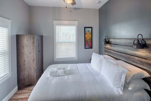 A bedroom with a large bed, a wooden wardrobe, a window with blinds, and a wall-mounted lamp.