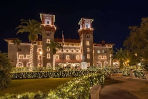 A large, historic building with two prominent towers, a well-maintained garden with lights, and a statue in front.