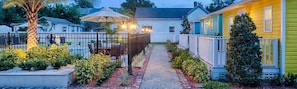 A pathway leading to a fenced patio area with a table and chairs, surrounded by greenery and a palm tree.