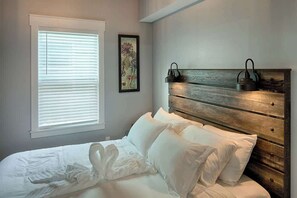 A bedroom with a wooden headboard, white bedding, and a window with blinds.