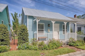 A light blue house with a porch and white railings, surrounded by a garden with various plants and flowers.