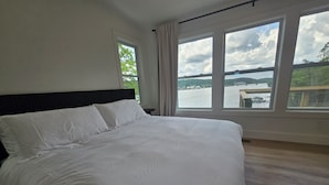 A large bed with white linens in a room with a view of a body of water and greenery.