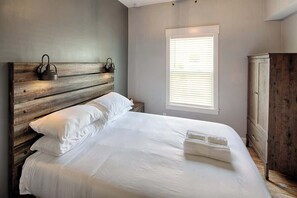 A bedroom with a wooden headboard, white bedding, a bedside lamp, and a window with blinds.
