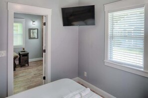 A bedroom with a bed, a TV mounted on the wall, a window with blinds, and a door leading to another room. - Charming St. Augustine cottages + resort-style outdoor area + walk to downtown. (St. Augustine)
