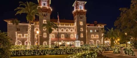 A large, historic building with two prominent towers, a well-maintained garden with lights, and a statue in front.
