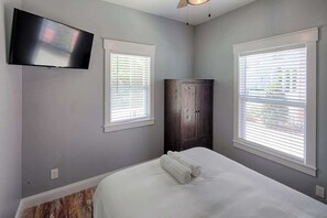 A bedroom with a bed, a flat-screen TV mounted on the wall, and two windows with blinds.