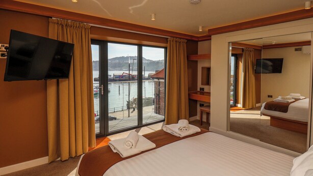 A hotel room with a bed, a TV, a balcony with a view of the water and buildings, and a mirror on the wall. - Apartment 2 (Scarborough)