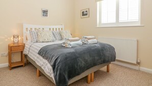 A bedroom with a bed, bedside table, lamp, and a window with blinds. - Malting Cottage (Dawlish)