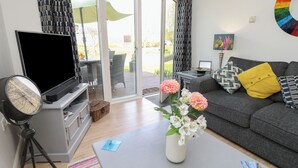 A living room with a flat-screen TV, a vintage-style globe, a coffee table with a vase of flowers, a grey sofa with patterned cushions, and a sliding glass door leading to an outdoor area.