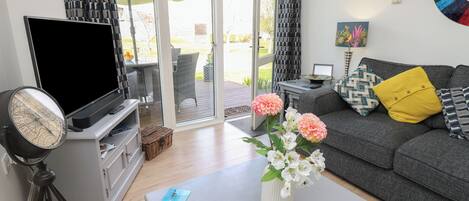 A living room with a flat-screen TV, a vintage-style globe, a coffee table with a vase of flowers, a grey sofa with patterned cushions, and a sliding glass door leading to an outdoor area.