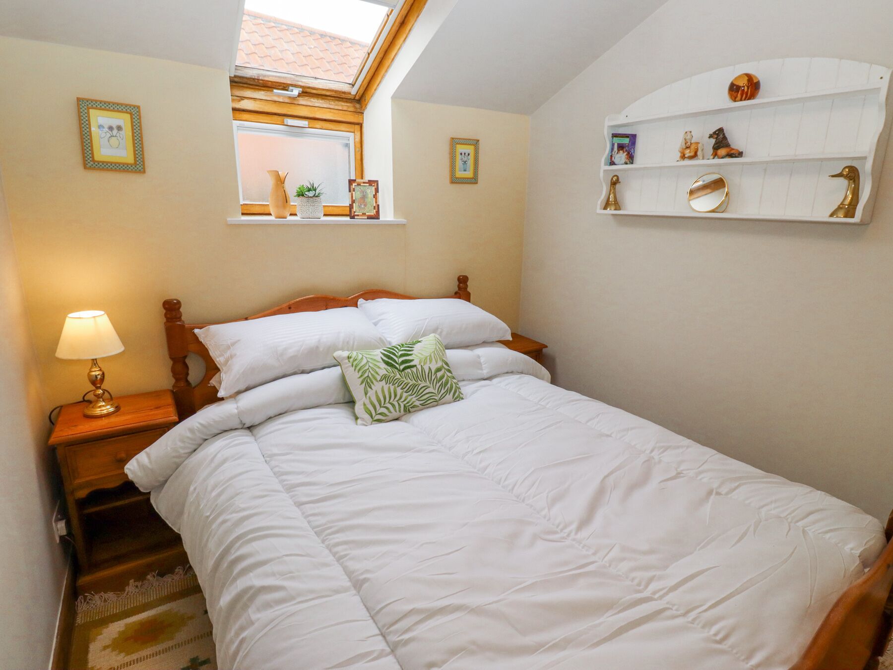 Una cama bien hecha con ropa de cama blanca, un cojín verde y un cabecero de madera. Hay una mesita de noche con una lámpara, una ventana con vistas al exterior y una estantería con objetos decorativos.