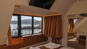A hotel room with a large window offering a view of a harbor, a bed with white linens, and a small seating area with a table. - Apartment 3 (Scarborough)