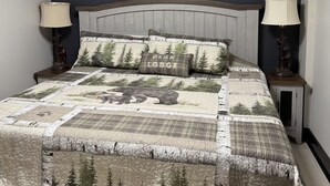 A bedroom with a bed featuring a forest-themed quilt, two bedside lamps, a headboard, and a skylight.