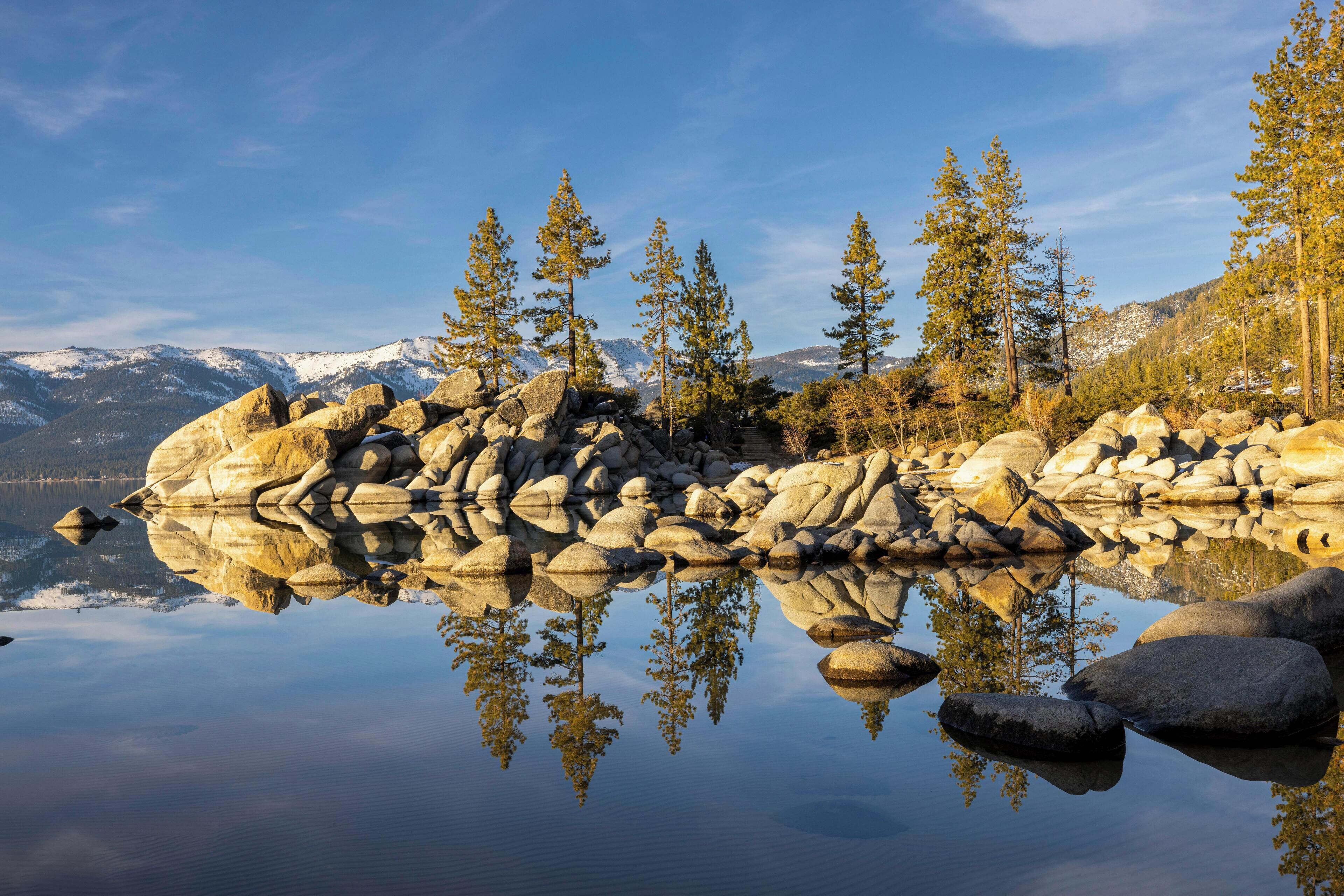 Un lac paisible aux eaux claires, où se reflètent les arbres et les rochers, entouré de montagnes et surplombé d’un ciel dégagé.