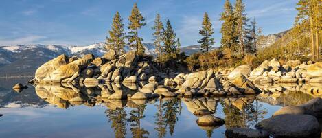 Un lac paisible aux eaux claires, où se reflètent les arbres et les rochers, entouré de montagnes et surplombé d’un ciel dégagé.