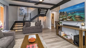 Uma sala de estar moderna com escada, sofá cinza, mesa de centro de madeira e uma pintura grande de paisagem costeira.