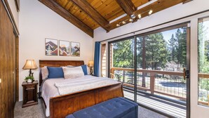 A bedroom with a large bed, a wooden headboard, a bench, a nightstand, and a view of a forest through a sliding glass door.
