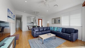 Ein modernes Wohnzimmer mit einem dunkelblauen Ledersofa, einem Holztisch und einem gemusterten Teppich. Ein Essbereich mit Tisch und Stühlen ist ebenfalls vorhanden, und im Hintergrund befindet sich eine Küche.