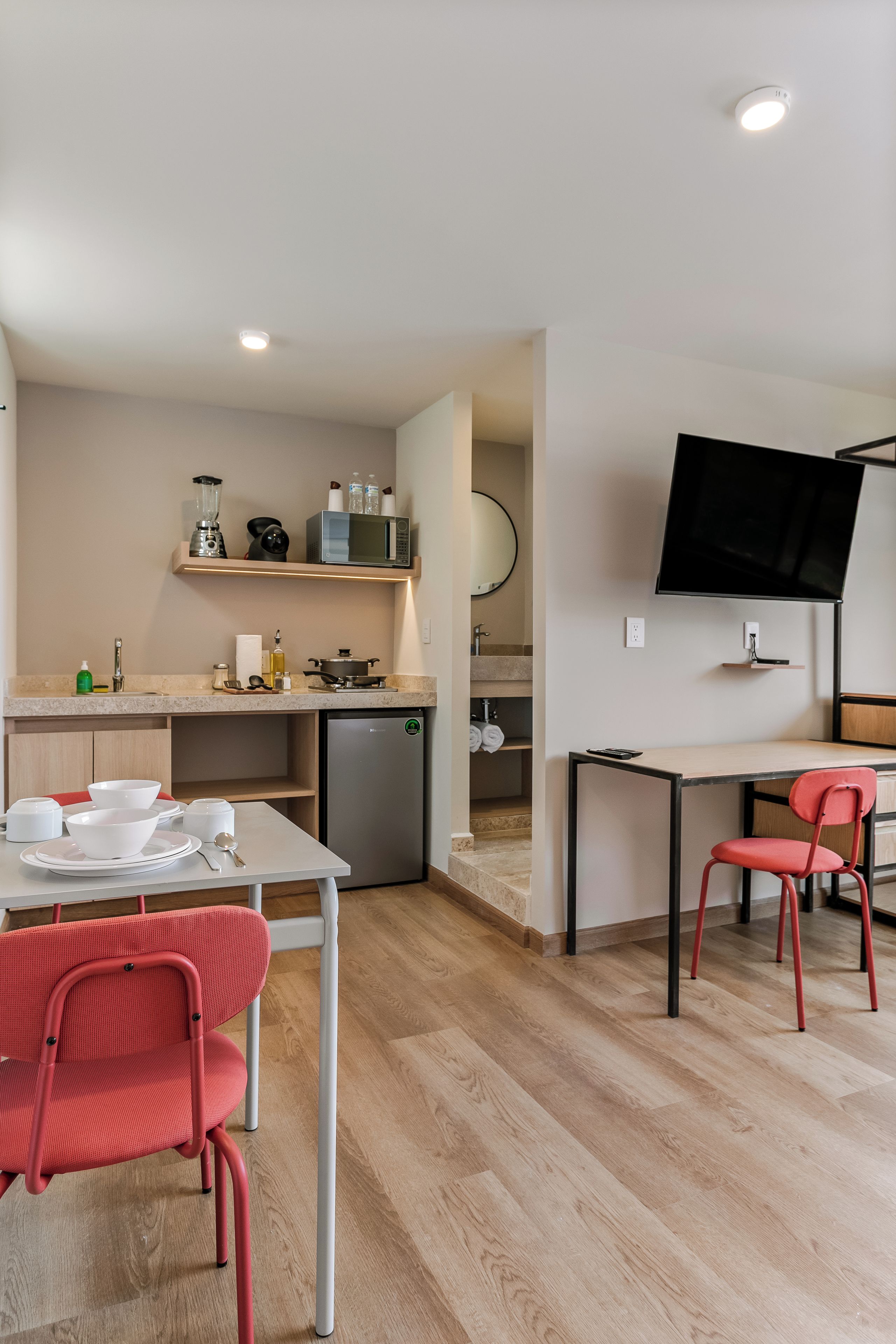 Cocina compacta con refrigerador, microondas y lavamanos. Hay una mesa de comedor con sillas y un televisor de pantalla plana montado en la pared.
