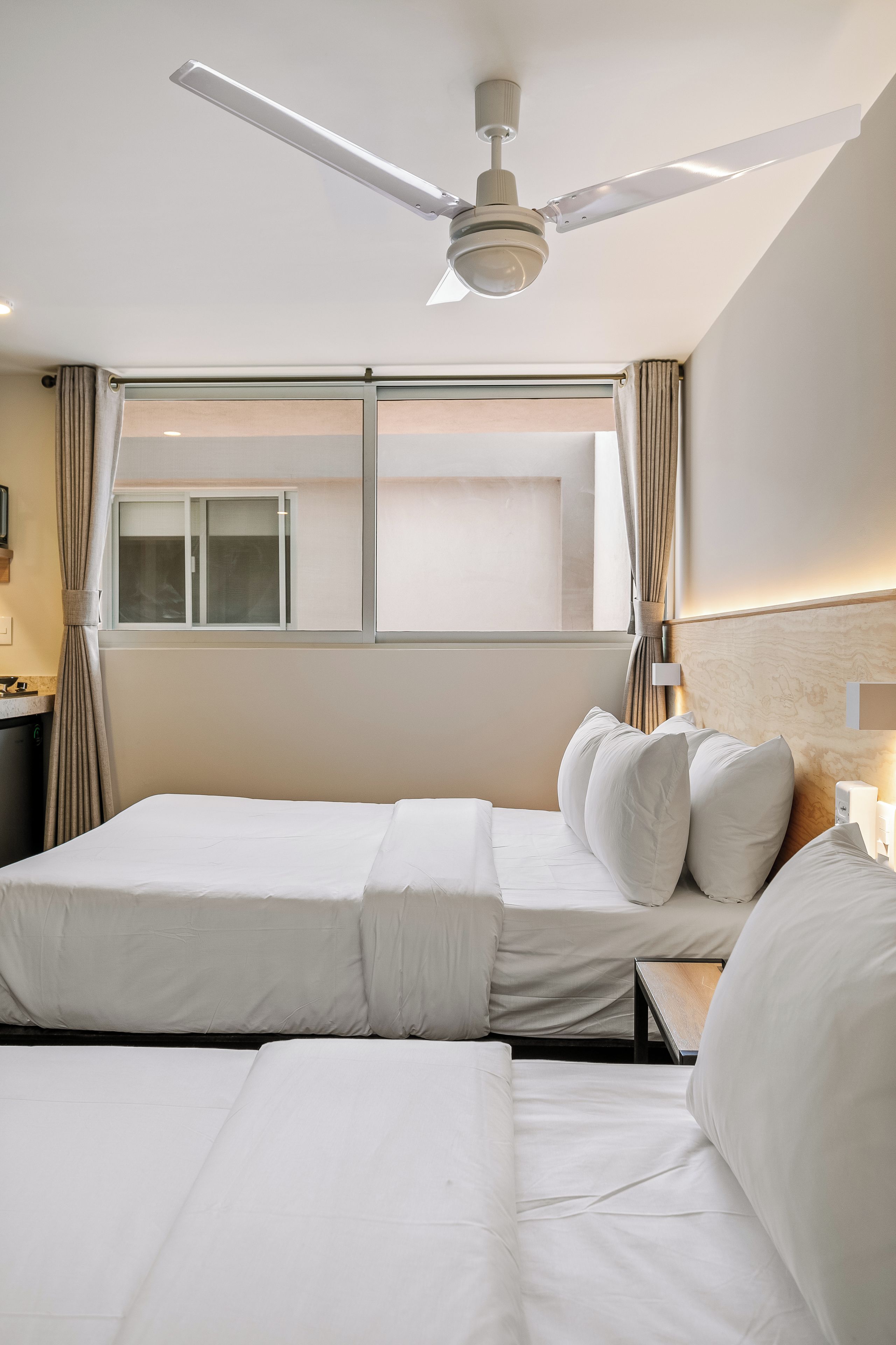 A hotel room with two beds, a ceiling fan, a window with curtains, and a nightstand with a lamp.