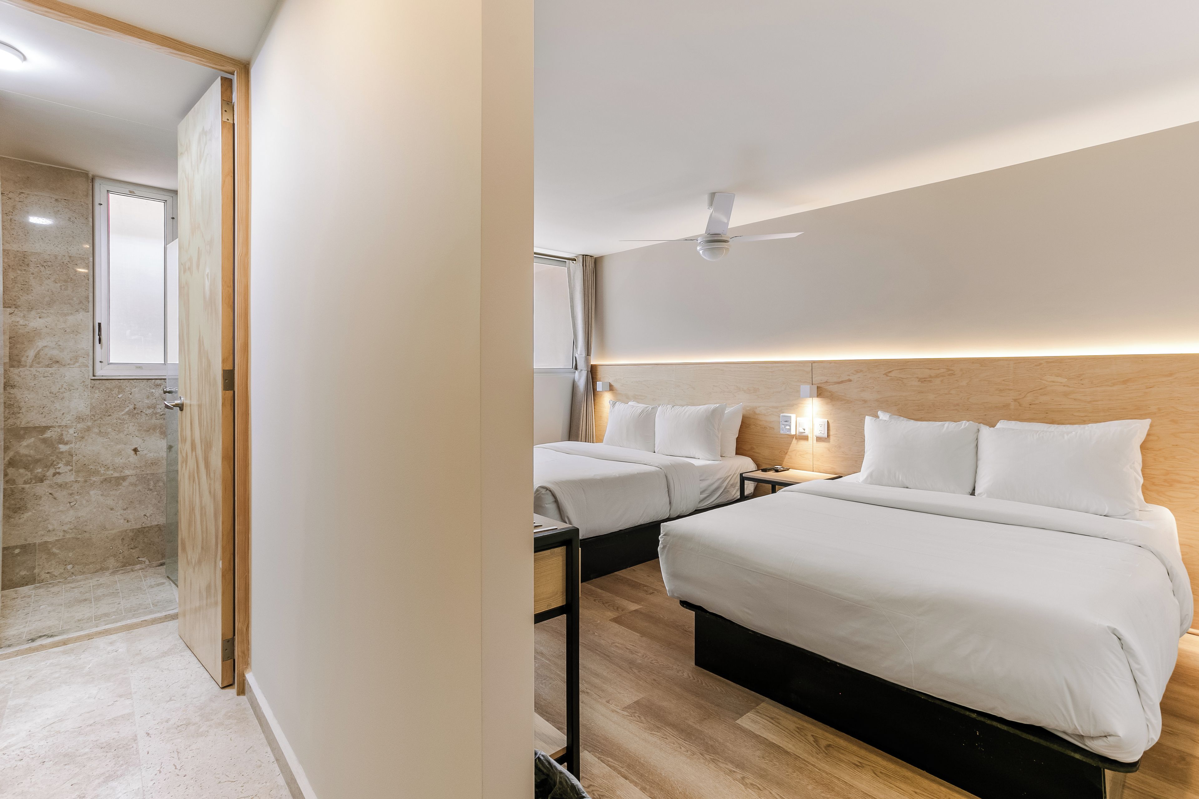 Une chambre d’hôtel avec deux lits, une tête de lit en bois, un ventilateur de plafond et une salle de bain avec une douche et un miroir.
