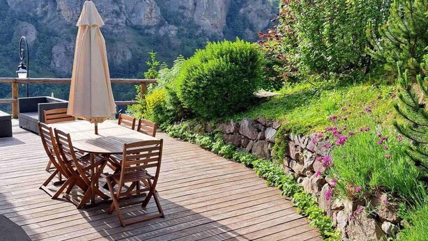 Una terraza de madera con una mesa y sillas, rodeada de follaje y un muro de piedra.