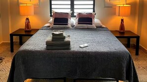 Una cama bien tendida con una colcha oscura, dos almohadas y una pila de toallas dobladas. Hay dos mesitas de noche con lámparas, un ventanal con persianas y dos cuadros en las paredes.