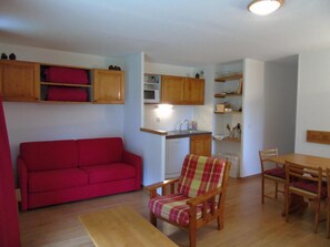 Een gezellige, kleine leefruimte met een rode bank, een ruitpatroon fauteuil, een houten eettafel en een kitchenette met een magnetron en spoelbak.