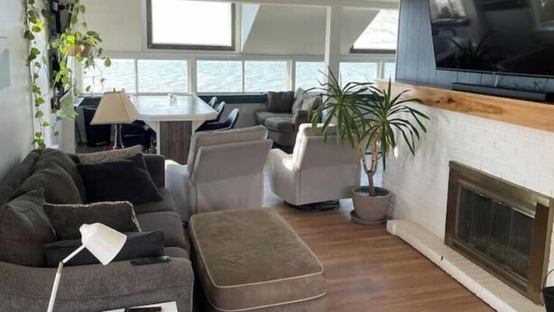 A modern living room with a sofa, ottoman, and a dining area with a table and chairs. There is a fireplace and a large TV mounted on the wall. - Lakefront Home Huron/Vermillion Amazing View (Vermilion)