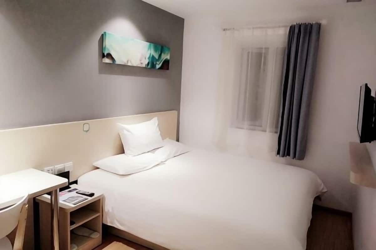A hotel room with a bed, a desk, a chair, a nightstand, a painting on the wall, and a window with curtains.