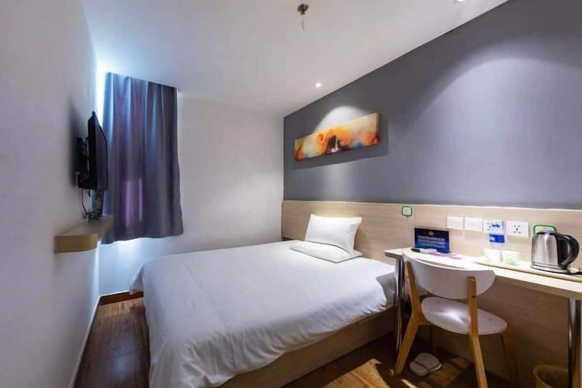 A hotel room with a bed, a desk with a chair, a painting on the wall, and a television on the wall.